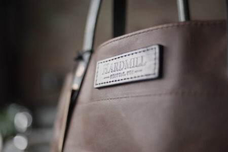 Close-up of a brown leather bag with a label that reads "Hardmill." The label is sewn onto the leather surface, which is smooth and well-crafted. The bag features metal rivets and dark handles.
