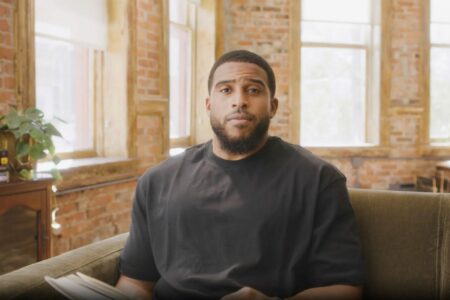 A man with a beard sits on a green couch in a bright room with exposed brick walls and large windows. He is holding a book, and a stack of books is visible on a wooden cabinet nearby.