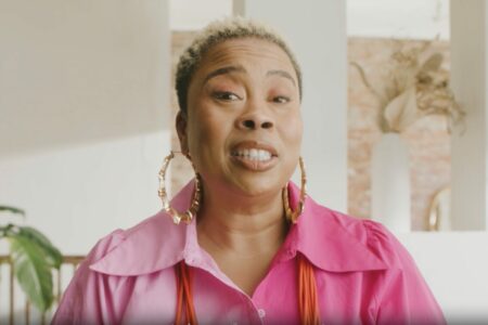 A person with short hair is smiling, wearing a bright pink shirt and large hoop earrings, standing indoors with soft natural light. The background features plants and a decorative arrangement on a shelf.