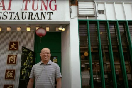 A person stands smiling in front of the entrance to Tai Tung Restaurant, which has a green door and red lanterns. Signs with Chinese characters are displayed next to the entrance.