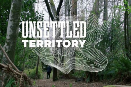 A dense forest with tall trees and green foliage. A person walks on a path in the background. The text 