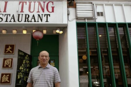 A man in a striped shirt stands in front of Tai Tung Restaurant. The sign above him displays the restaurant's name. A red lantern hangs near the entrance, and tall green bars are visible on the adjacent wall.
