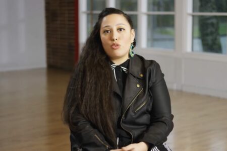 A person with long hair sits in a bright room, wearing a black leather jacket and statement earrings. A large window reveals a glimpse of greenery outside. The person appears to be engaged in conversation.