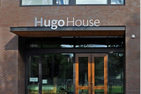 Brick building with large windows displaying "Hugo House" above glass doors. The entrance includes wood-framed doors, and the building number "1640" is visible.