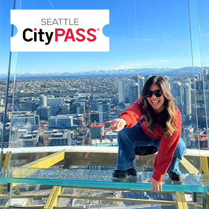 A person in a red sweater and sunglasses crouches and points on a glass platform high above a cityscape. The Seattle CityPASS logo is in the top left corner.