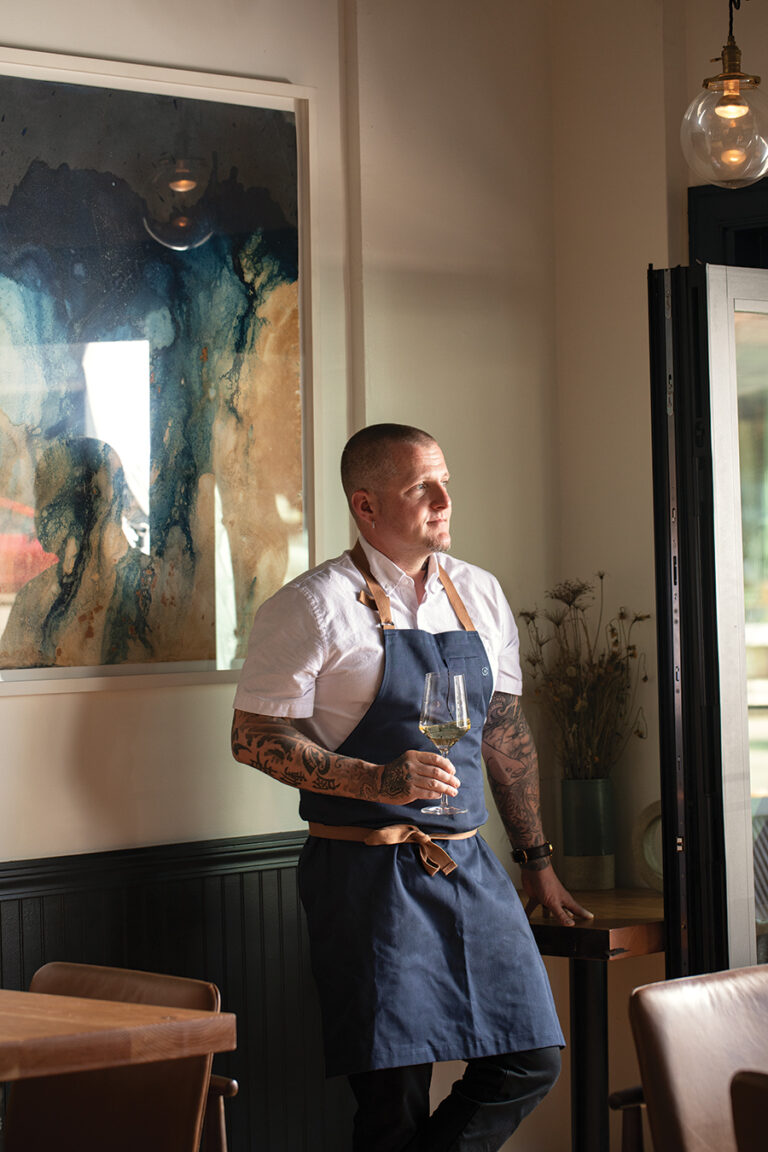 A person wearing a white shirt and blue apron stands in a stylish room holding a wine glass. They have tattoos on their arms and are looking out of the window thoughtfully. The wall features a blue and beige abstract painting.