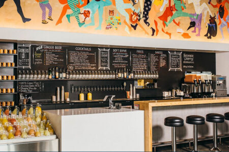 A modern bar with a colorful mural above. The bar features a variety of drinks, with bottles and glasses neatly arranged. A blackboard displays the menu with options for cocktails, soft serve, fries, and snacks. Five stools are at the counter.