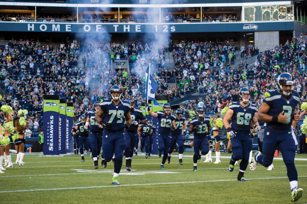 A football team charges onto the field, surrounded by cheerleaders and a roaring crowd in a packed stadium. Smoke fills the air, while a large banner overhead reads "HOME OF THE 12s," capturing the spirit of Seattle neighborhoods like SoDo and Georgetown.