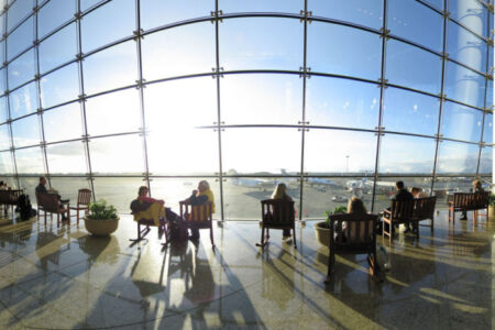 People seated in a spacious airport terminal, facing large windows with a view of the tarmac. The sun is shining brightly outside, casting long shadows on the floor. Airplanes and clear sky are visible in the background.