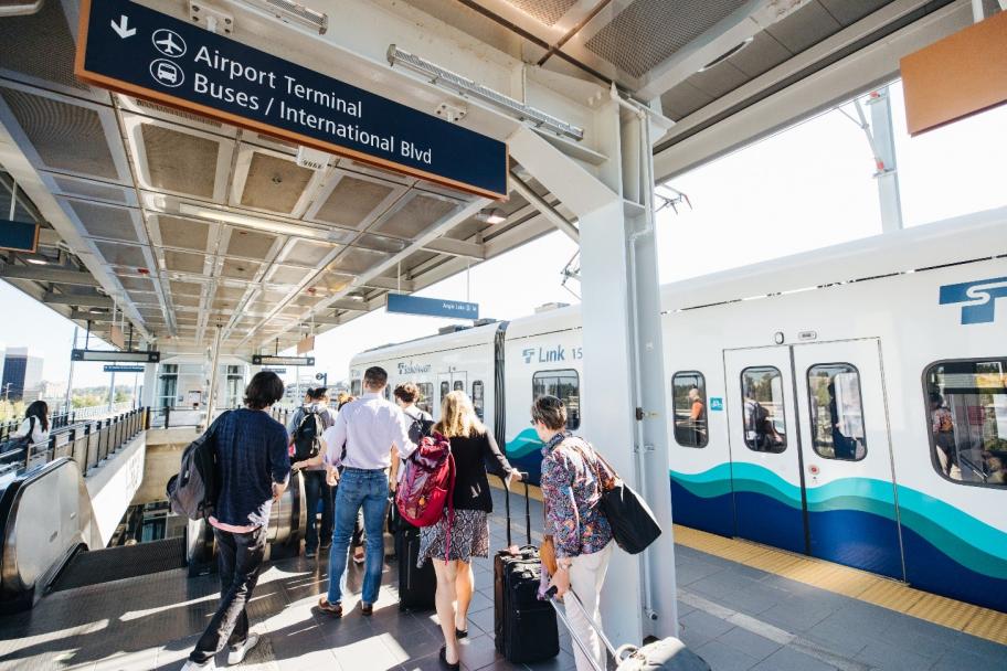People with luggage wait on a train platform next to a light rail train. An overhead sign directs to the airport terminal, buses, and International Blvd. The platform is bright and modern, with clear daylight outside.