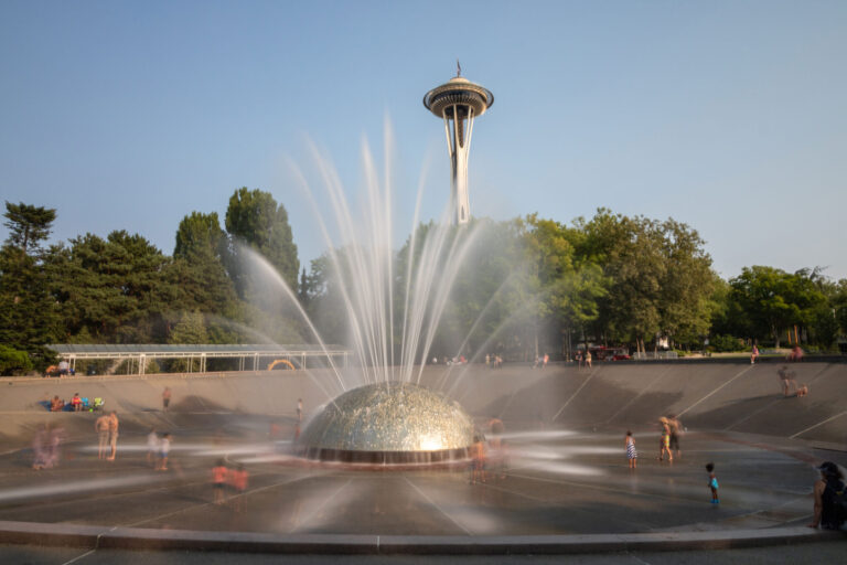 Children play around the dome-shaped International Fountain in Seattle, a highlight among the many things to do in Seattle. It sprays water in various patterns as the iconic Space Needle rises prominently in the background, surrounded by lush green trees under a clear blue sky.