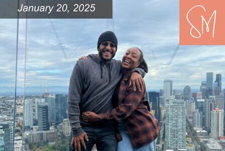 A smiling couple embraces on a rooftop with a city skyline in the background.