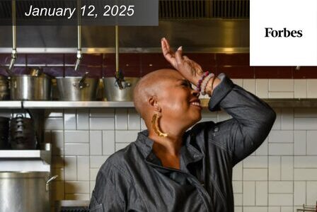 Chef Kristi Brown smiling in a kitchen setting, wearing a black jacket and large earrings, with a hand raised near their face.