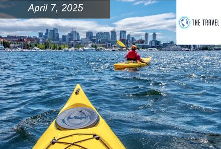 Two people kayaking on a blue lake with a city skyline in the background.