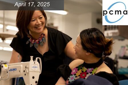 Two women smile and talk beside a sewing machine in a workspace.