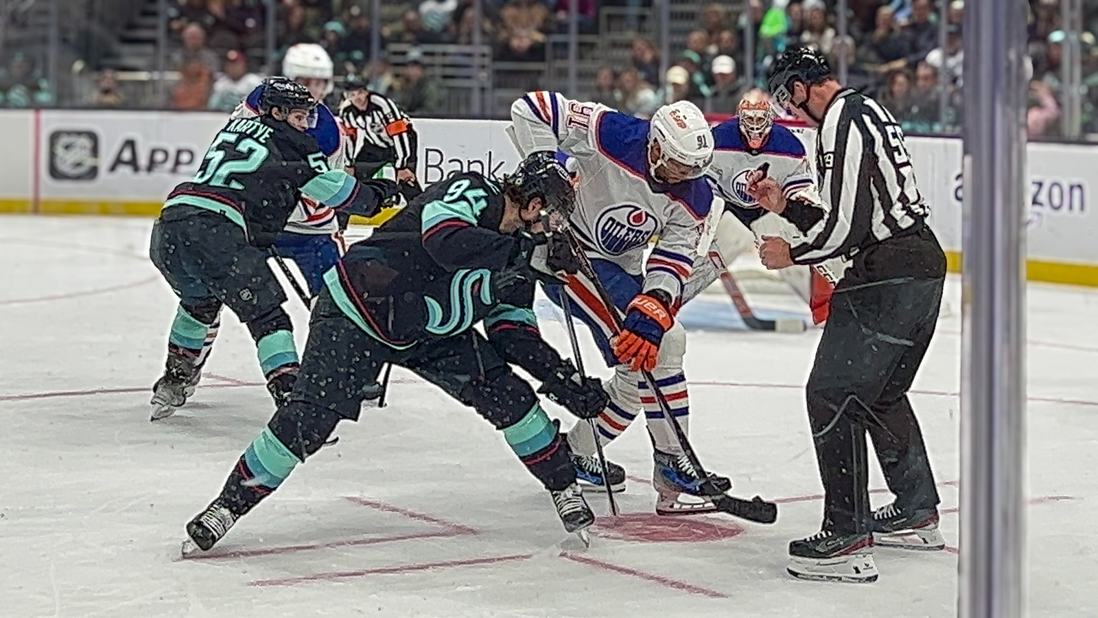 A Seattle Kraken player in the navy and aqua uniform goes up against the opposing team wearing a red, white, and blue uniform. They play on an ice rink with red markings. The ref stands to the right of the match up in a black and white uniform.