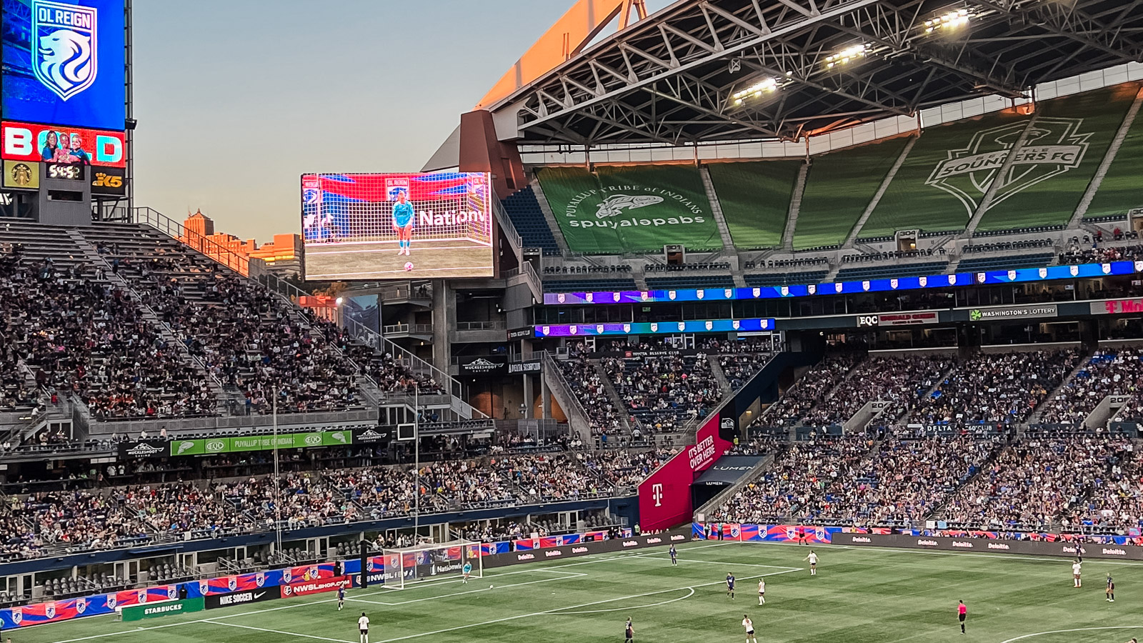Reign FC playing a match at Lumen Field. The sky is casting a glowy light onto the green turf field. A large TV screen is in the left center of the image, highlighting the keeper on the field who is about to take a kick.