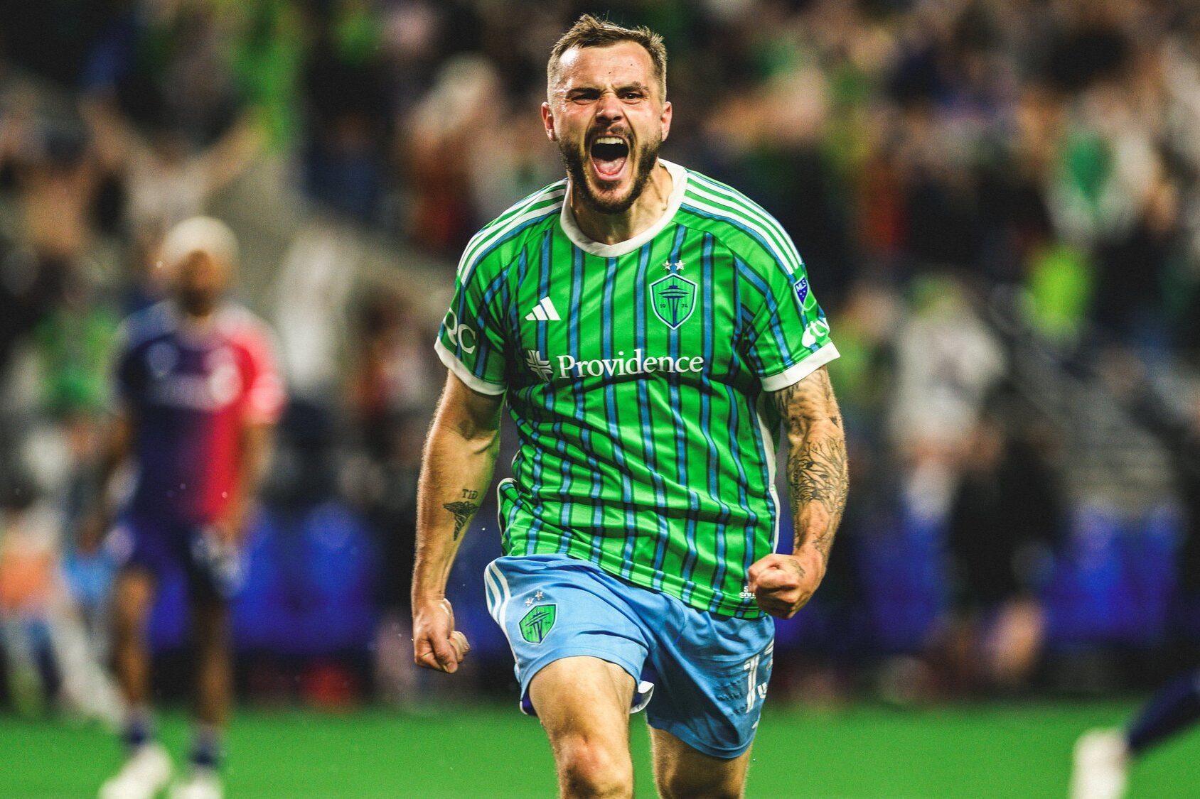 A Seattle sports player in a green and blue striped jersey is celebrating passionately on the field. He has his fist clenched and is mid-shout, with blurred crowds in the background.