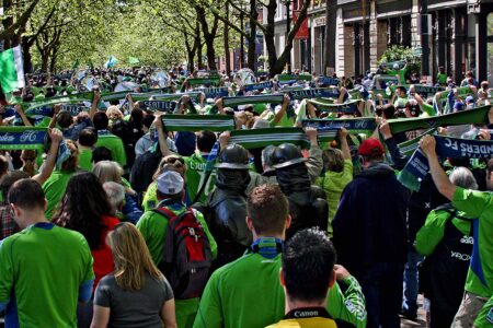 A large crowd of people, mostly wearing green and blue, gather on a tree-lined street. Many hold up scarves reading 