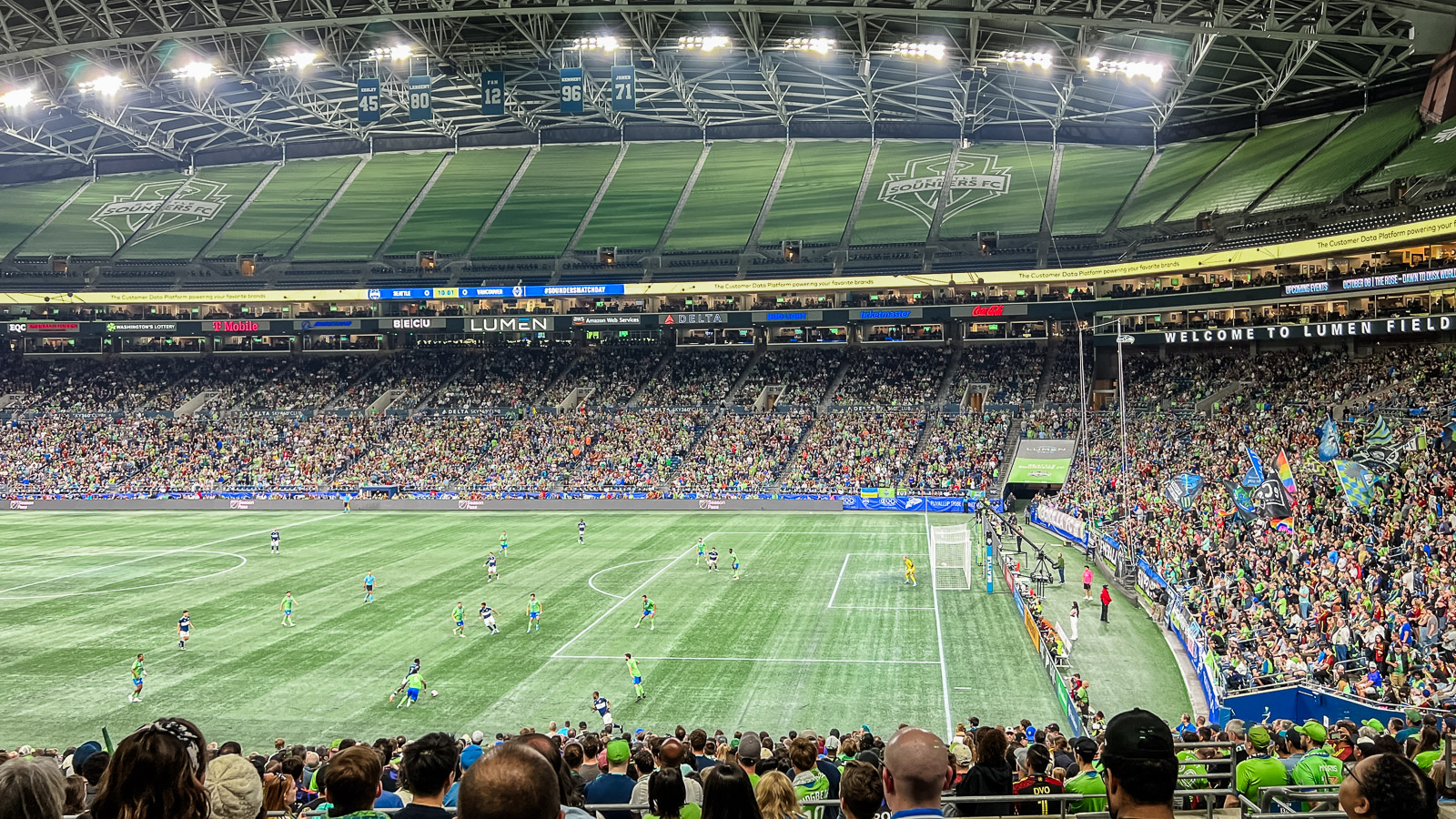 The Seattle Sounders soccer team and opponents on the bright green turf pitch at Lumen Field. Thousands of fans fill the lower bowl of the stadium. The top level has a green graphic cover with the Sounders logo in white. Fans on the far right wave colorful flags.