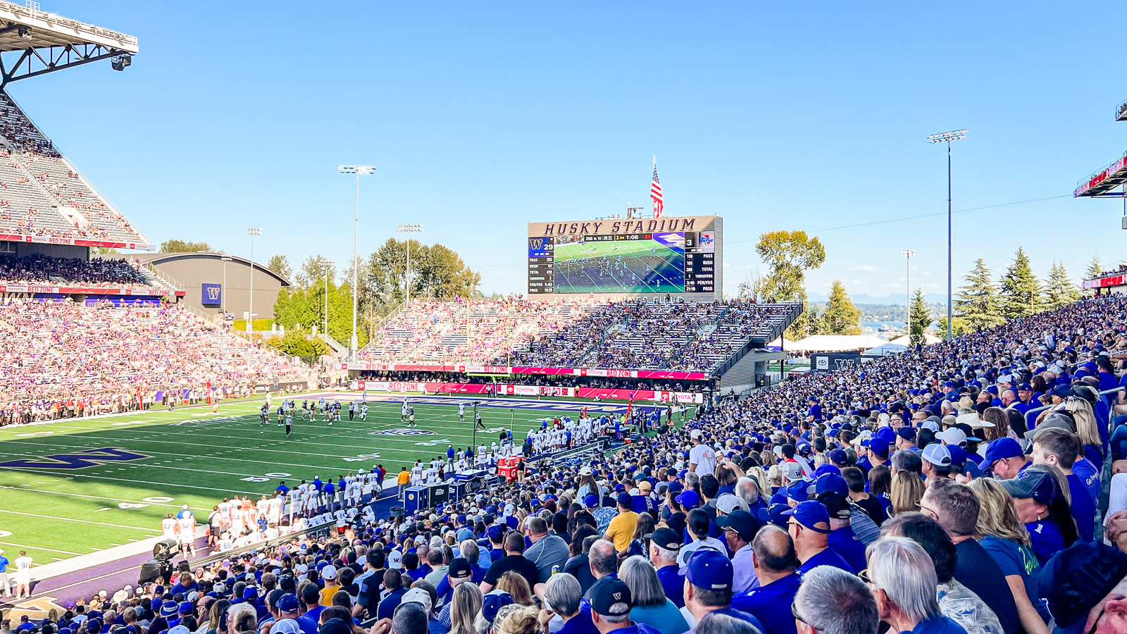 A sea of fans wearing purple and gold fill the the seats at Huskry Stadium while watching the school's football team on the bright green field.