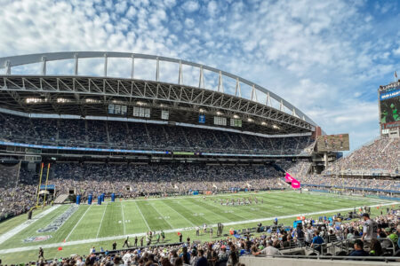 Thousands of fans fill the seats at Lumen Field on a sunny day in Seattle. The bright green turf with players in the center of the field fills the middle of the image.