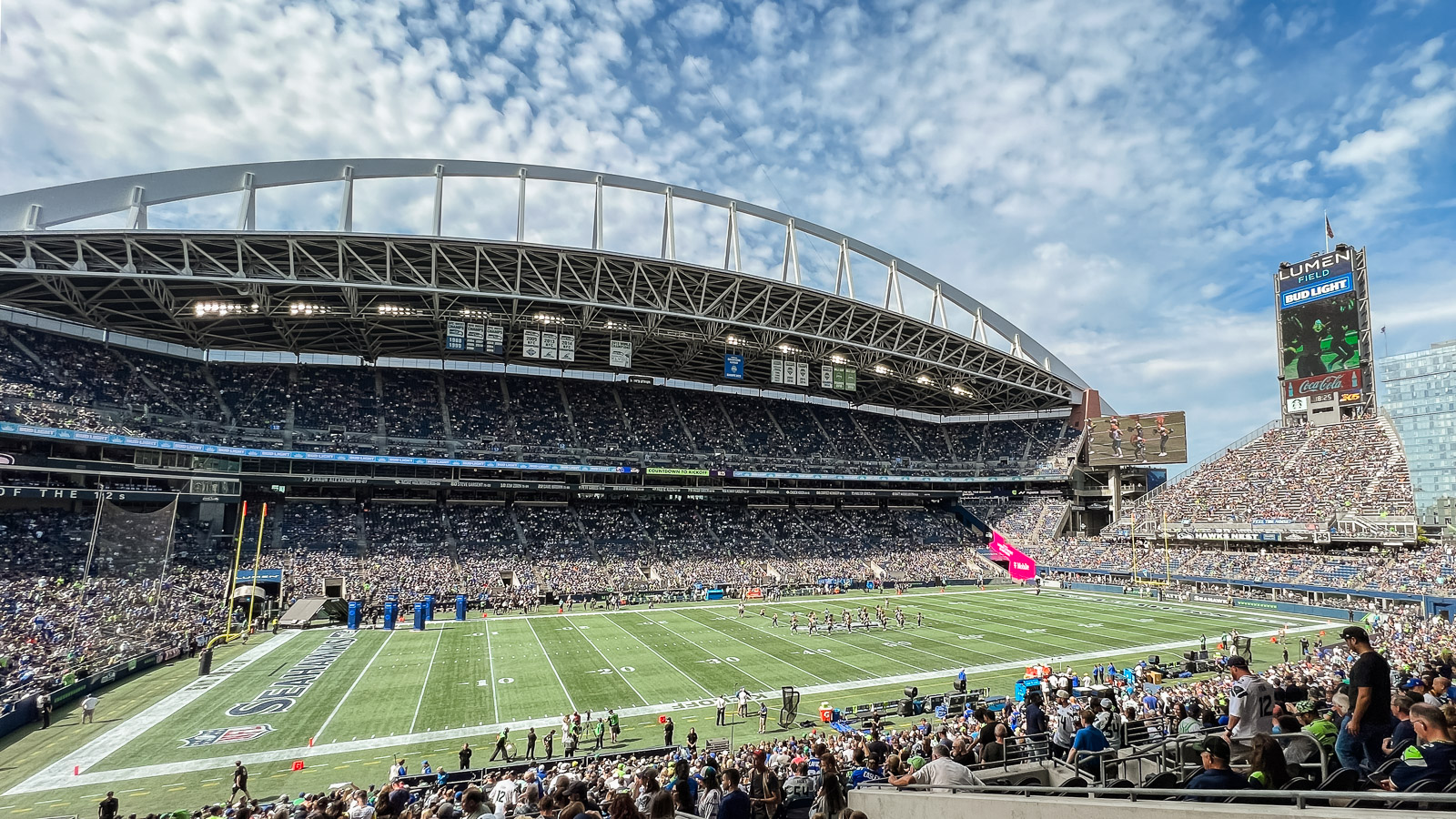 Thousands of fans fill the seats at Lumen Field on a sunny day in Seattle. The bright green turf with players in the center of the field fills the middle of the image.