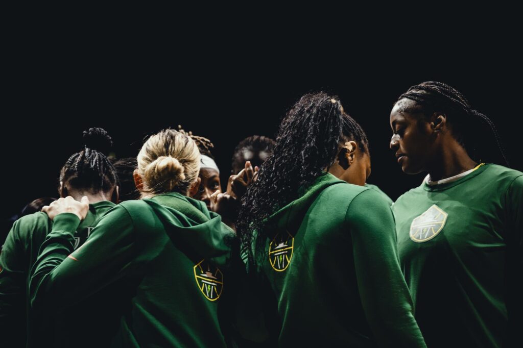 A group of basketball players in green uniforms huddled together, facing inward. The dimly lit scene captures the spirit of Seattle sports, emphasizing teamwork and unity. Their uniforms proudly display a yellow emblem on the back, symbolizing their unyielding bond.