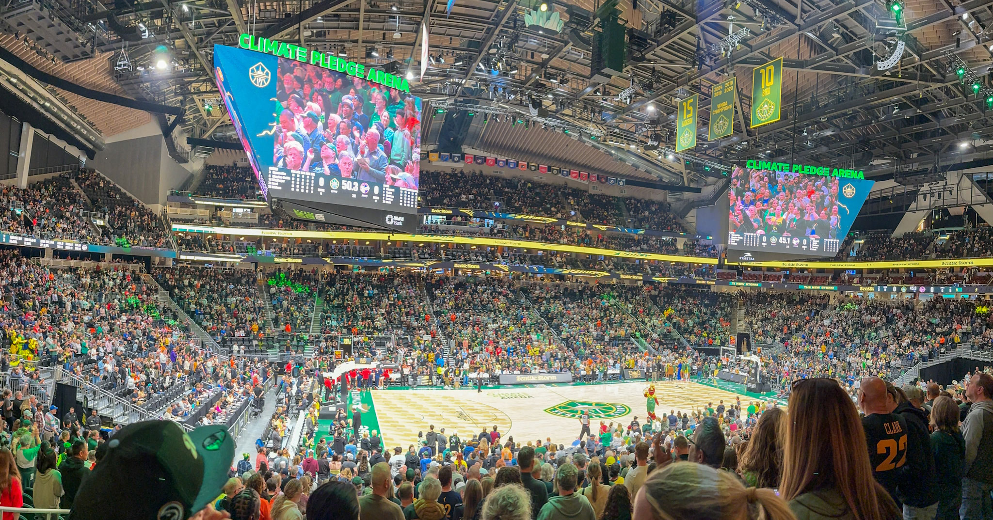 Climate Pledge Arena packed with fans watching the Seattle Storm game. Two giant TV screens float above the court, showing fans in the arena.