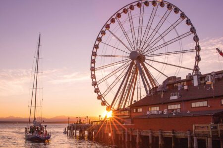 A scenic view of a Seattle waterfront neighborhood during sunset showcases a large Ferris wheel on the pier, a graceful sailing boat gliding through the water, and an iconic building. The sky boasts a blend of orange and pink hues as the sun peeks through the Ferris wheel.