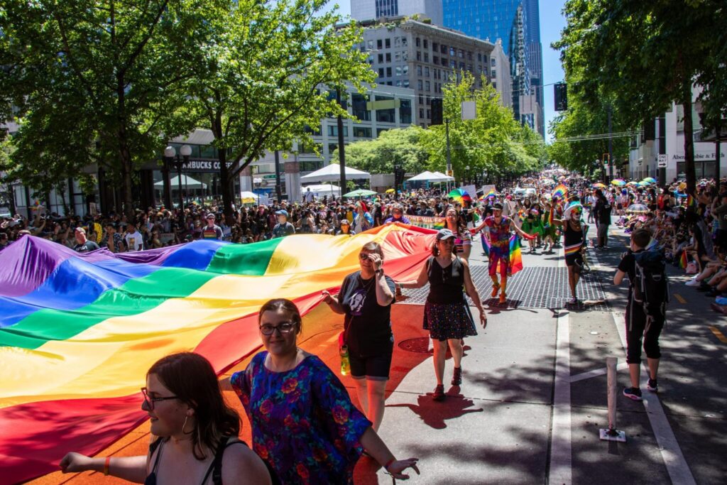 A large group of people march in a city parade, carrying a giant rainbow Pride flag. Spectators line the street, and trees provide shade on a sunny day. Many participants wear colorful outfits and wave smaller rainbow flags.