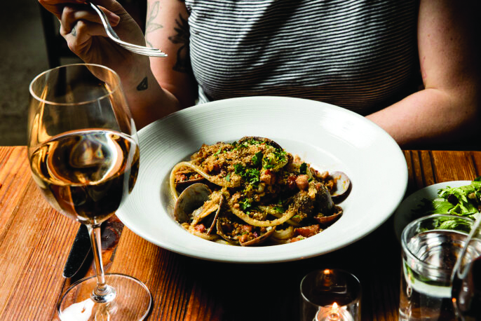 A person in a grey shirt sitting at a wood table with a white plate full of pasta and a glass of white wine.