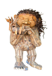 A large, whimsical sculpture of a creature made from various pieces of wood, featuring exaggerated facial features, thick hair made from twigs, and holding a smaller wooden figure. The sculpture is detailed and textured.