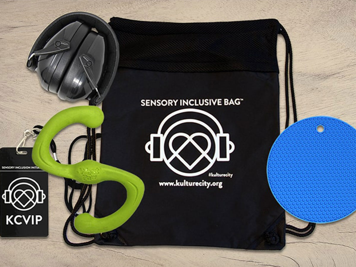 A sensory inclusive bag featuring items: noise-canceling headphones, a KultureCity ID, a green sensory toy, and a blue textured disc, with the logo and website "kulturecity.org" displayed on the bag.