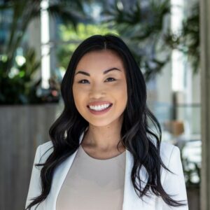 A person with long dark hair, wearing a white blazer and beige top, smiles at the camera. The background features indoor plants and modern decor, suggesting a professional setting.