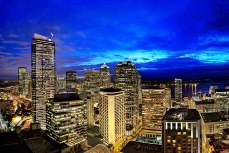 A nighttime cityscape featuring illuminated skyscrapers under a vibrant blue sky. The tall buildings are brightly lit, contrasting against the dark evening. In the background, faint hints of water and distant lights are visible.