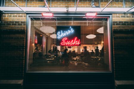 A restaurant window at night with "Shiro's Sushi" in neon lights. Inside, people are seated at tables, enjoying their meals. The ambiance appears cozy and intimate, with soft lighting and a warm atmosphere.