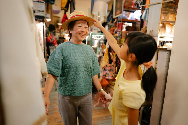 A woman wearing a green shirt smiles as a child in a yellow shirt puts a straw hat on her head. They're enjoying some family fun and exploring one of the bustling, colorful markets—a must-visit among things to do in Seattle—filled with bags and various treasures.