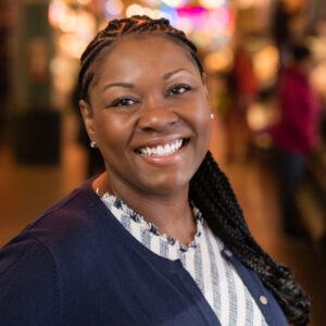 A person with braided hair smiles warmly in a bustling indoor setting. They are wearing a blue cardigan over a striped top, and colorful lights are blurred in the background, suggesting a lively atmosphere.