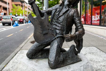 A bronze statue of a musician kneeling with an electric guitar is displayed outdoors on a stone pedestal. It's situated on a city sidewalk with buildings and trees in the background.