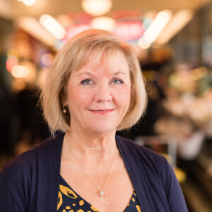 A woman with short blonde hair and a blue patterned blouse smiles at the camera. The background is softly blurred, suggesting a bustling indoor setting with warm lighting.