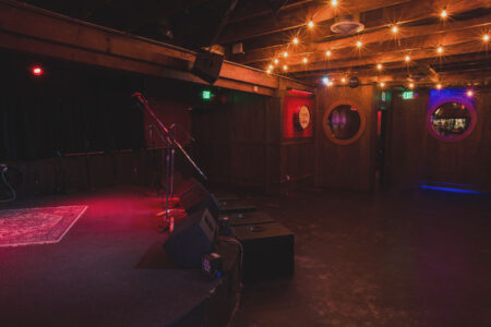 A dimly lit music venue with a stage, microphone stands, and an amplifier. The ceiling is adorned with string lights, creating a warm ambiance. Circular windows on dark wood walls provide a cozy, intimate setting.