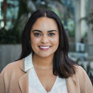 A person with long dark hair smiles warmly at the camera. They are wearing a white blouse and a beige coat. The background is softly blurred, with indoor plants and bright natural lighting creating a serene atmosphere.