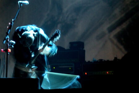 A musician on stage intensely bending forward while playing an electric guitar. The background is dimly lit, with amplifiers and equipment visible. The scene conveys motion and energy, highlighted by blurred effects.