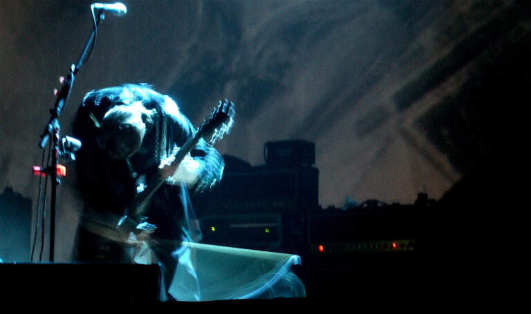 A musician on stage intensely bending forward while playing an electric guitar. The background is dimly lit, with amplifiers and equipment visible. The scene conveys motion and energy, highlighted by blurred effects.