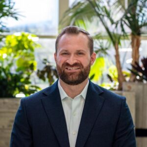A man with a beard, wearing a suit and a light-colored shirt, is smiling in an indoor setting with large windows. Lush green plants are visible in the background, creating a bright and natural atmosphere.