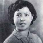 A vintage black and white portrait of a young woman with short, curly hair. She is smiling softly and wearing a collared, textured sweater. The background is softly blurred, adding to the timeless feel of the image.