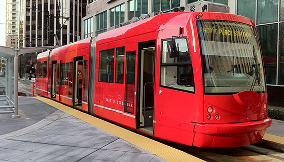 A red streetcar is stopped at a station on a city street. The digital display reads "Pacific Place Station." The surroundings include tall buildings and a sidewalk with a yellow tactile paving strip.
