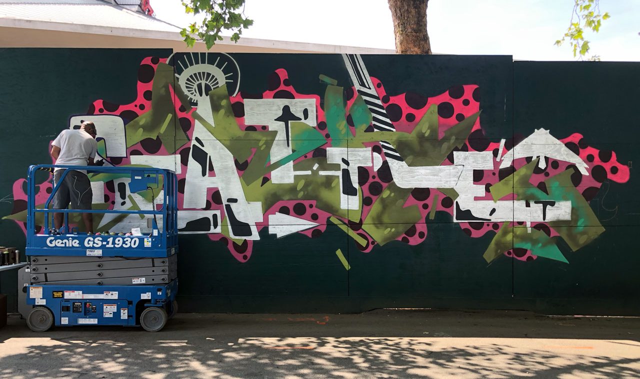 A person on a blue Genie lift paints a colorful mural on a large wall. The artwork features bold, abstract shapes in green, pink, black, and white. A tree with green leaves stands in the background, partially shading the wall.