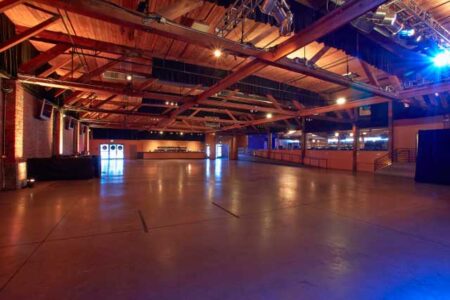 A spacious, empty warehouse venue with a wooden ceiling and structural beams. The polished concrete floor is unobstructed, and bright lights illuminate the space. Doors and a raised platform are visible in the background.